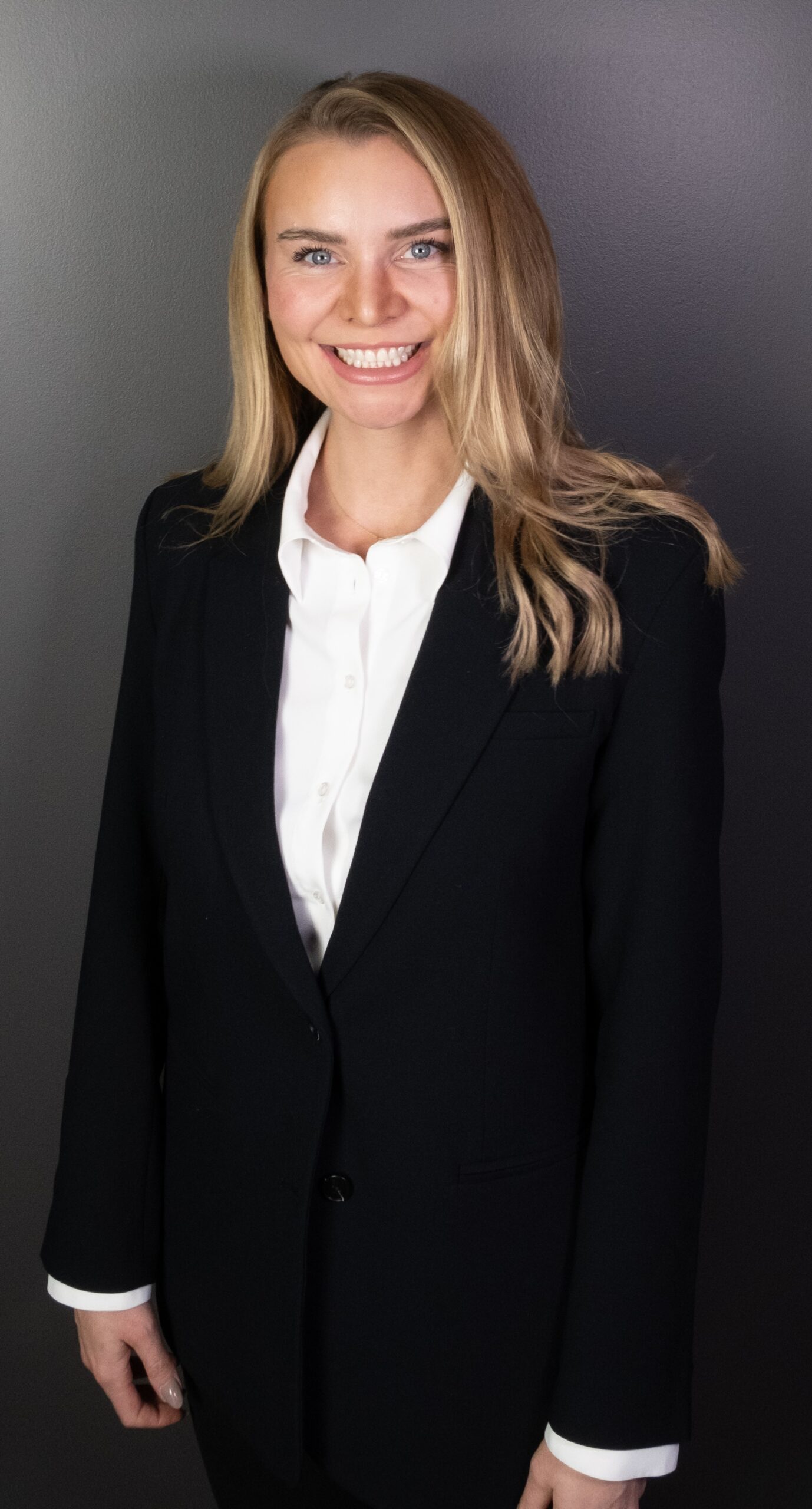 A professional woman in a black blazer and white shirt smiling confidently against a dark background.