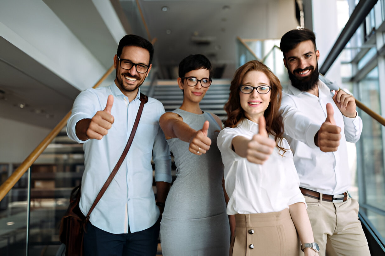 A group of four young professionals standing in an office building, smiling and giving a thumbs-up gesture.